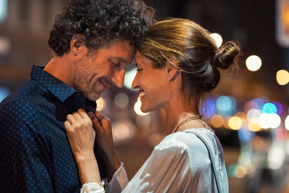 A couple shares a tender moment, smiling and leaning towards each other in a softly lit urban setting.