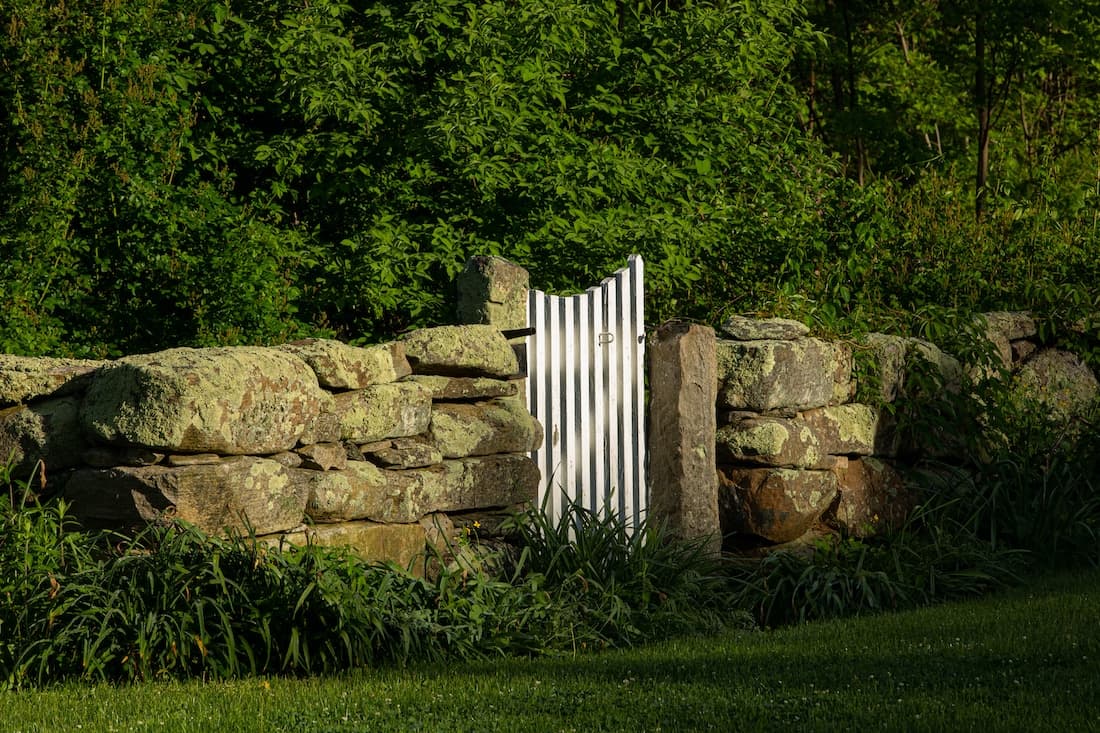 A white gate stands ajar in a stone wall surrounded by lush greenery.