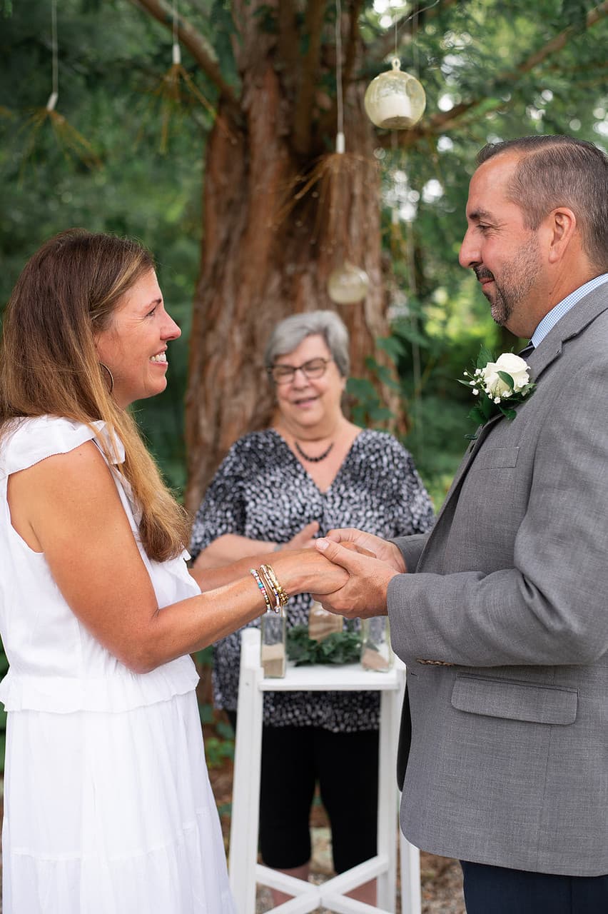 A couple exchanging vows during a wedding ceremony, with an officiant present in a lush outdoor setting.