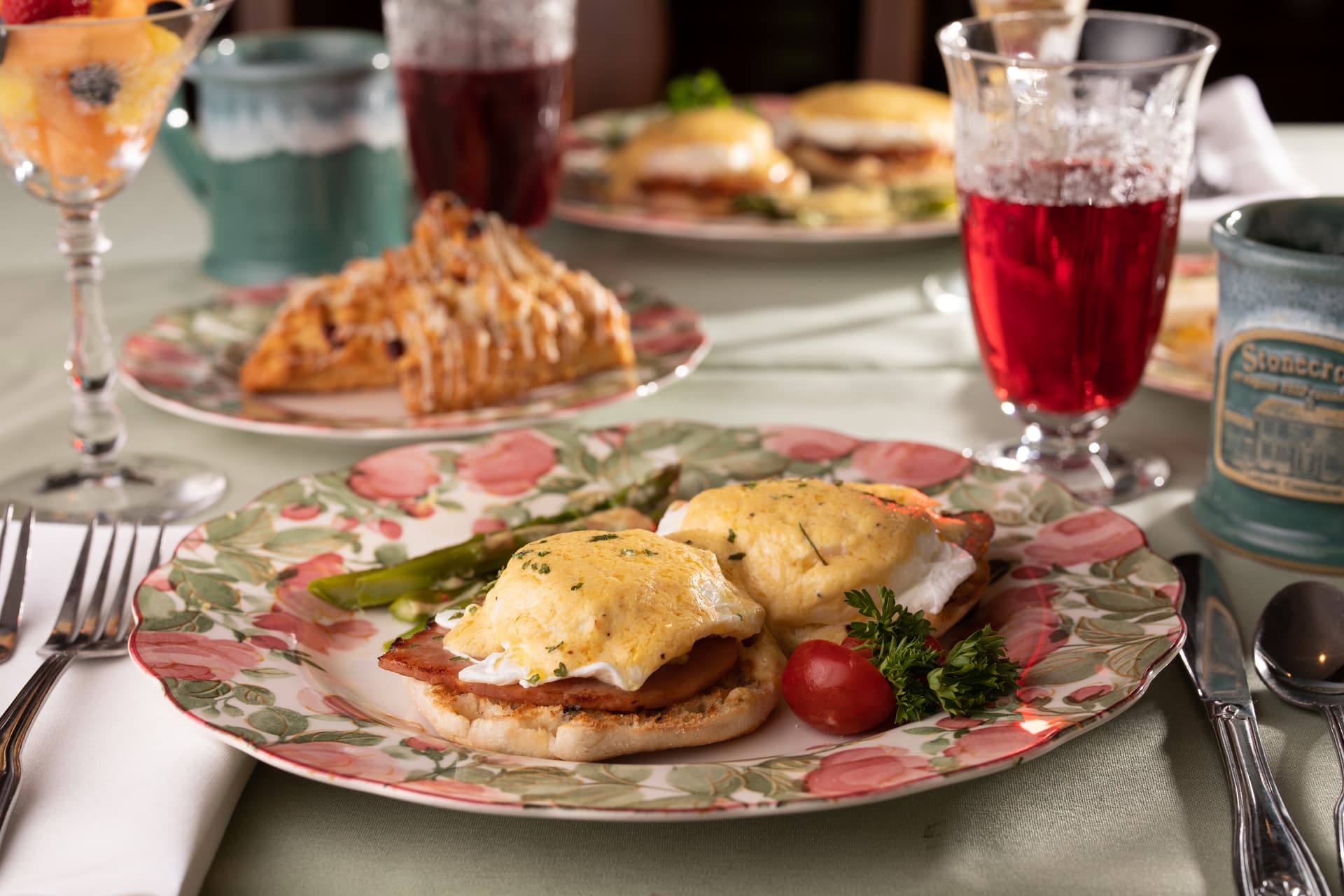 A plate of eggs Benedict with asparagus and tomato, surrounded by various breakfast items and drinks.