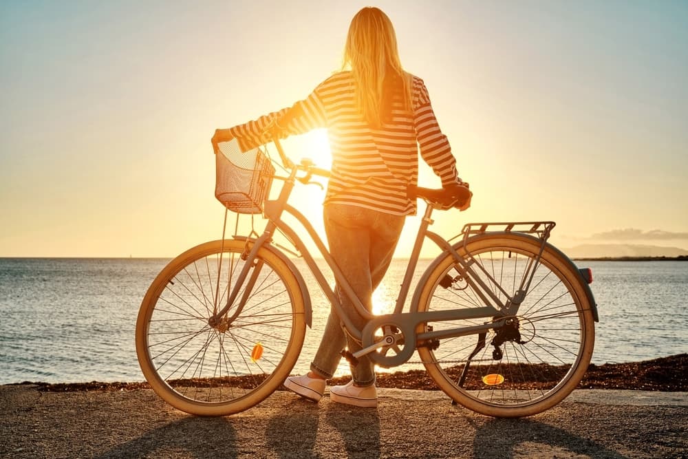A person stands beside a bicycle, silhouetted against a sunset over the water.