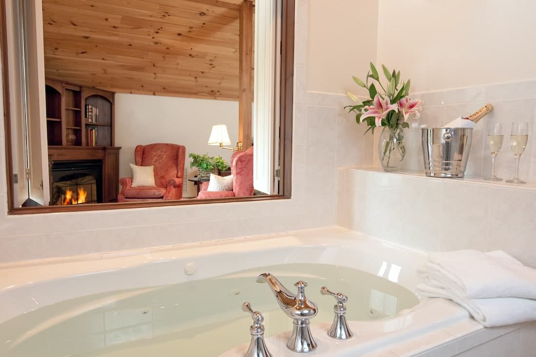 A luxurious bathroom featuring a whirlpool tub, a cozy living area visible through a window, and elegant decor including flowers and champagne.