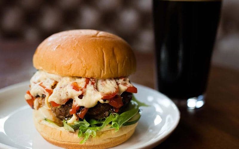A mouthwatering burger topped with cheese and sauce, served on a plate with a glass of dark beverage.