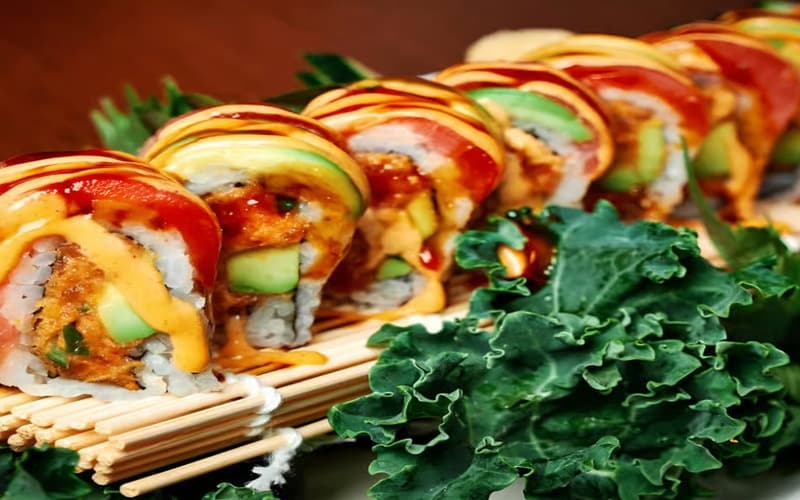 A colorful platter of sushi rolls topped with sauces and garnished with greens.