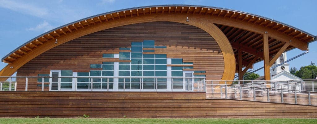 A modern wooden building with a curved roof and large glass windows, surrounded by green grass.