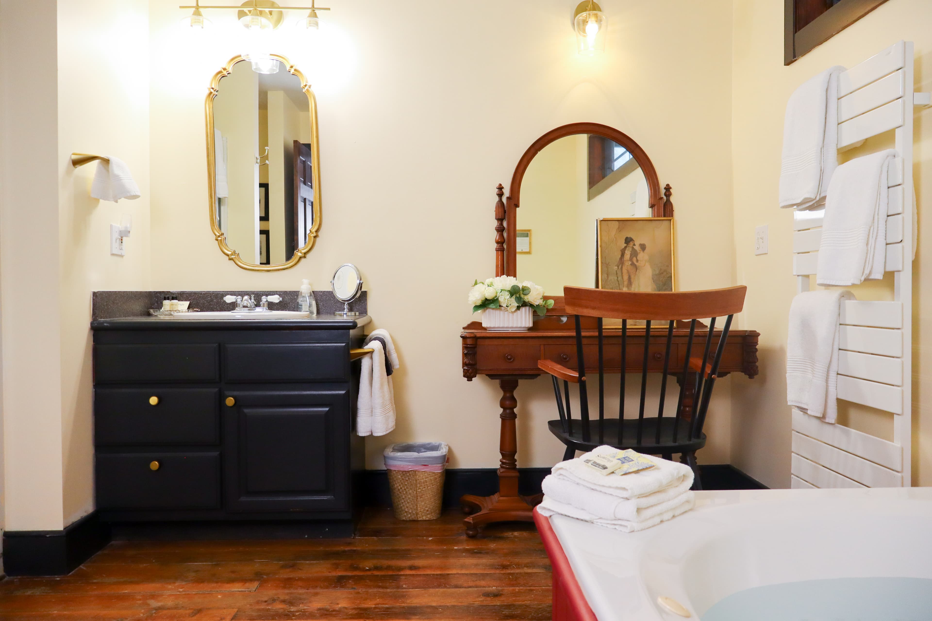 A bright bathroom featuring a dark vanity, a mirror, a wooden chair, and neatly stacked towels.