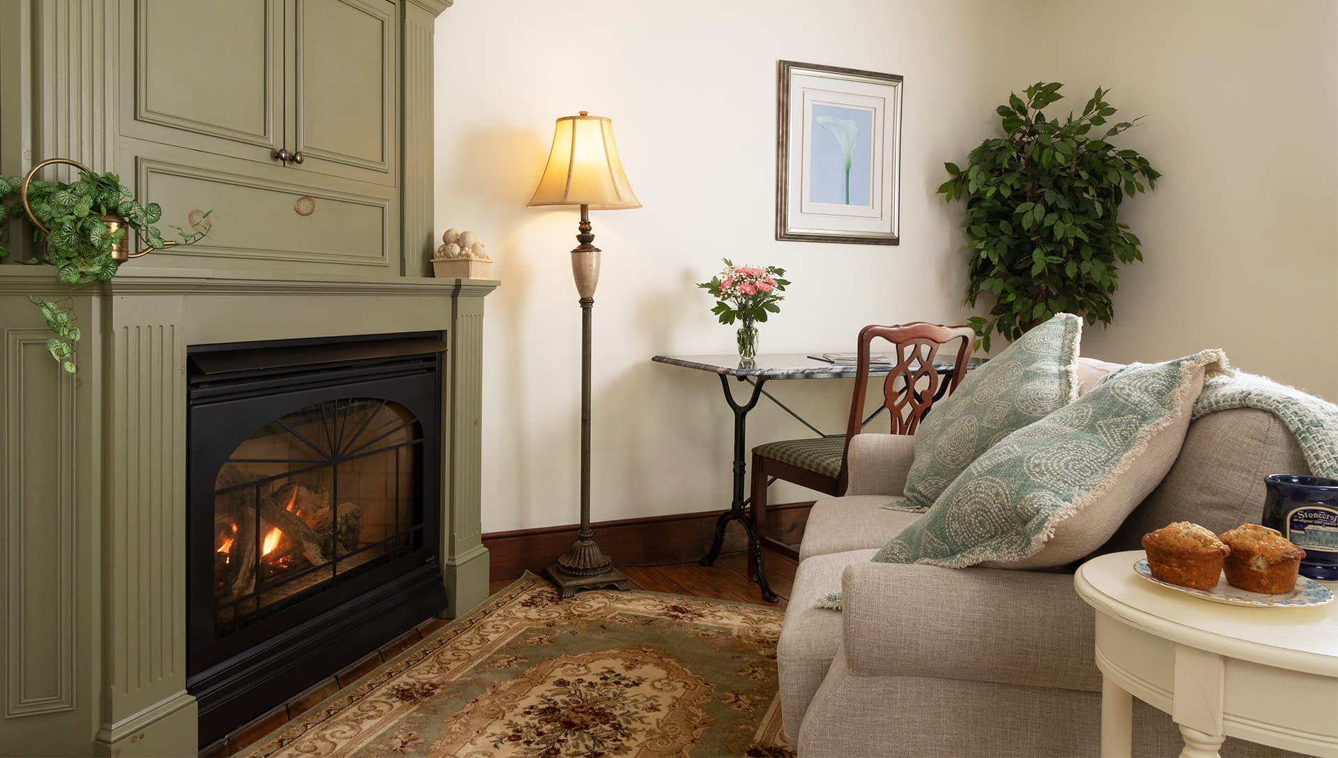 Cozy living room featuring a beige sofa, fireplace, lamp, and a small table with flowers and muffins.