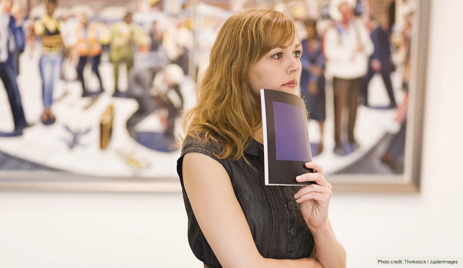 A woman stands thoughtfully in an art gallery, holding a brochure while observing a colorful mural.