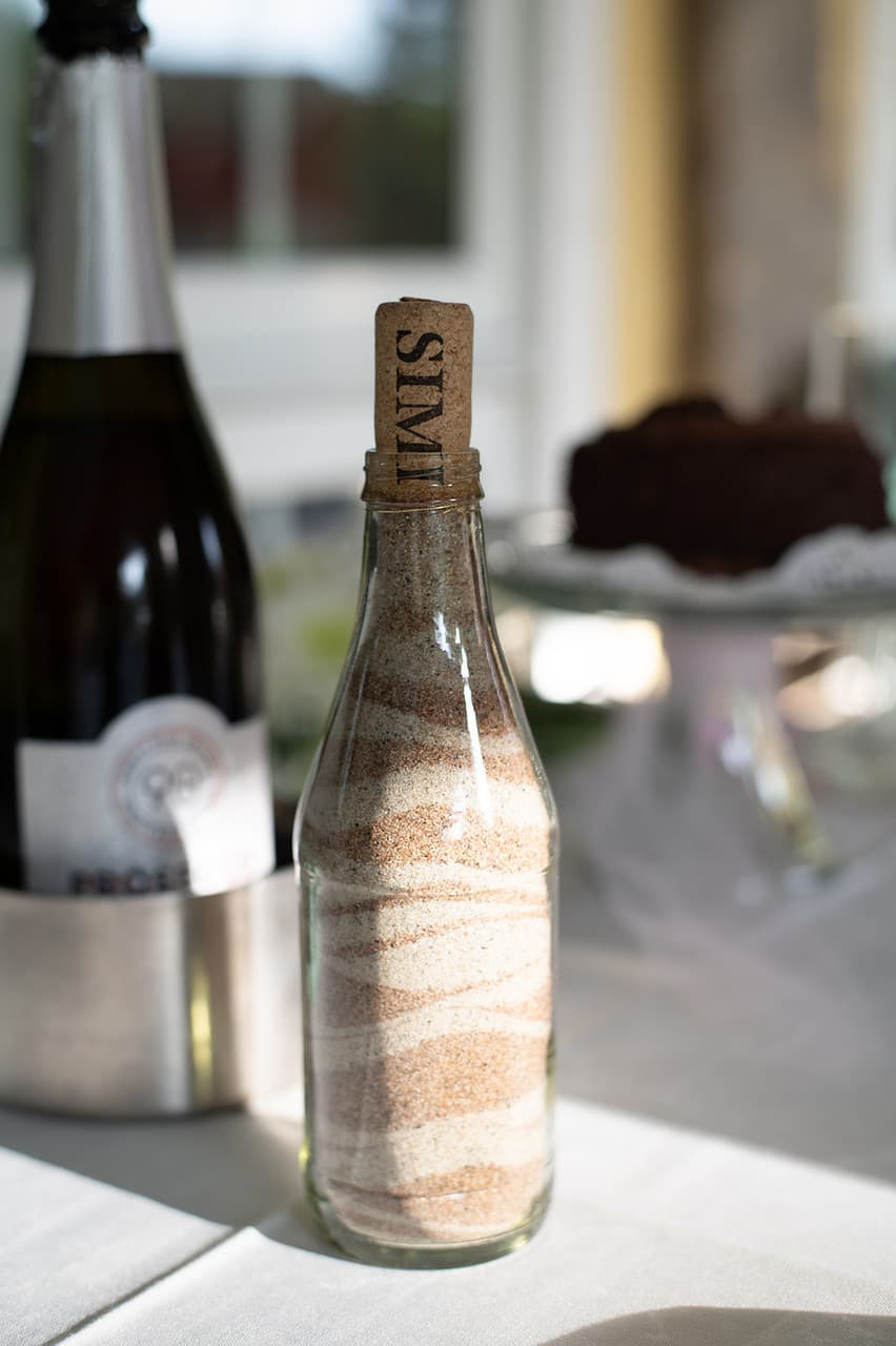 A glass bottle filled with layered sand and topped with a cork labeled "SALT" rests on a table beside a bottle of sparkling wine.