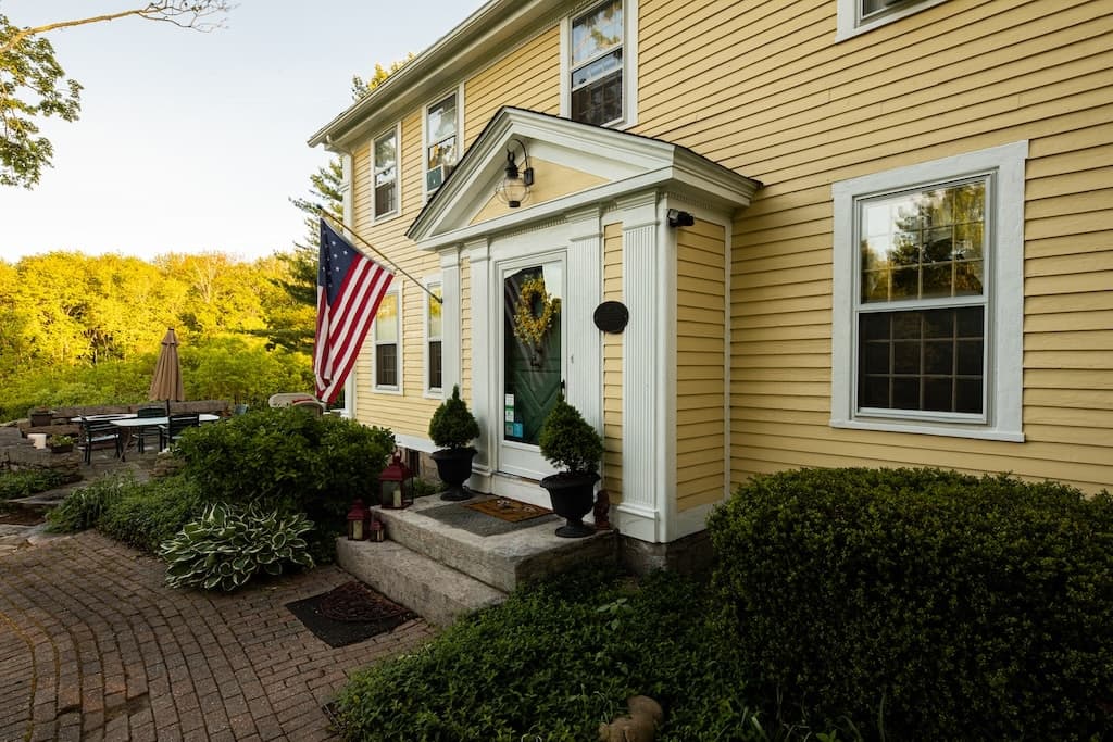 A yellow house entrance featuring a flag, potted plants, and a brick walkway.