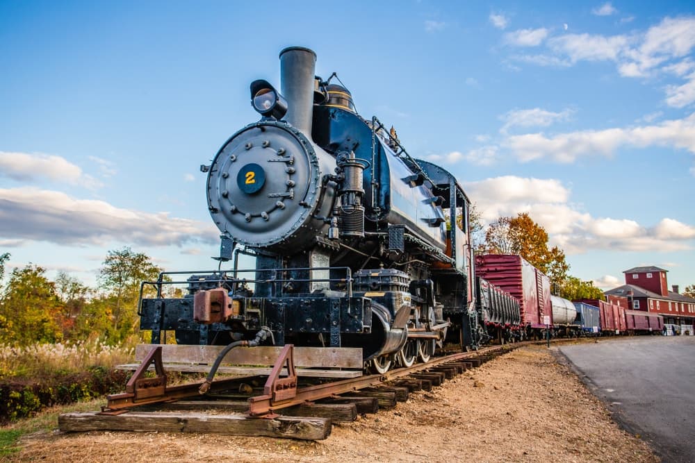 A vintage steam locomotive on tracks beside freight cars and autumn foliage.