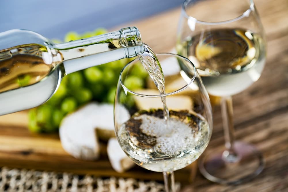 A bottle of white wine pouring into a glass, accompanied by cheese and grapes.