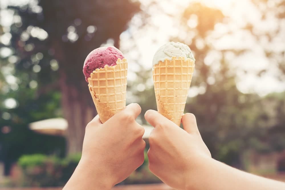 Two hands hold ice cream cones with pink and green scoops against a blurred natural background.