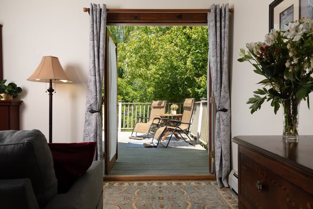 A view from a cozy room featuring open doors leading to a sunny deck with chairs and a serene garden outside.