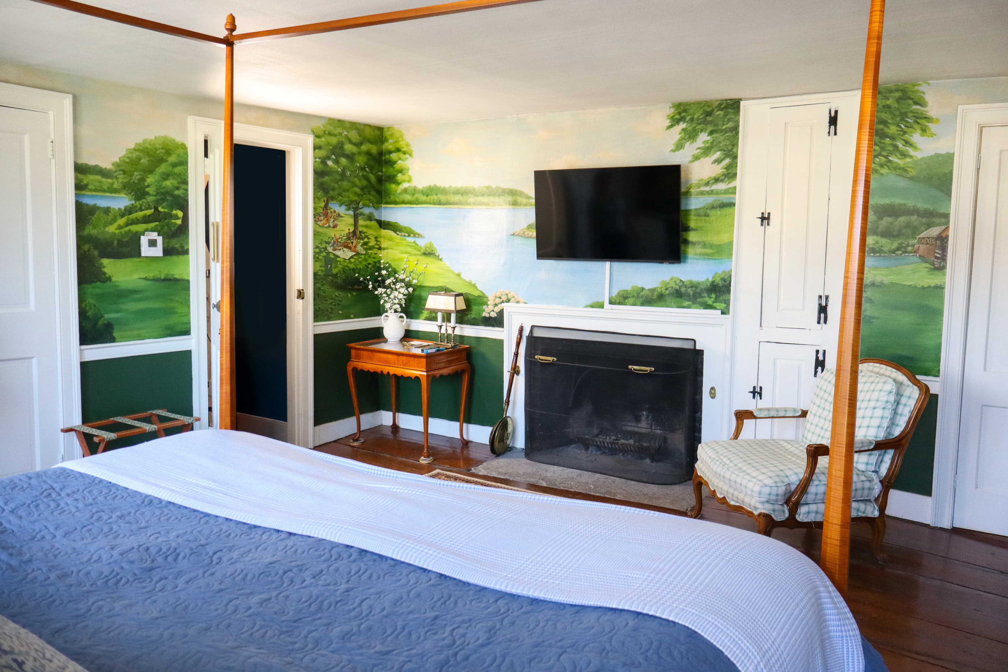 A cozy bedroom featuring a four-poster bed, a fireplace, and a scenic landscape mural.