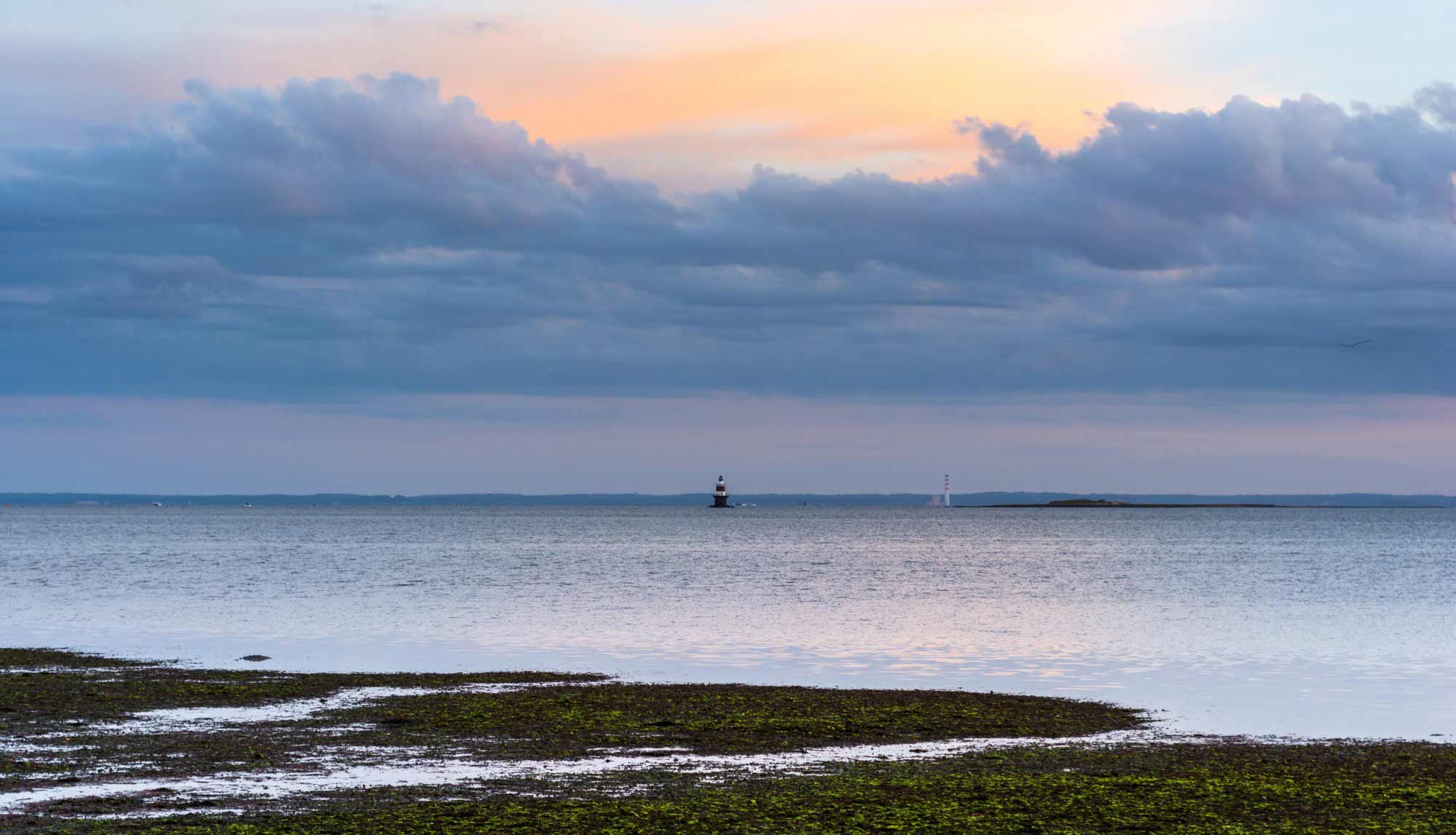 A calm sea at sunset with a lighthouse in the distance and clouds in the sky.