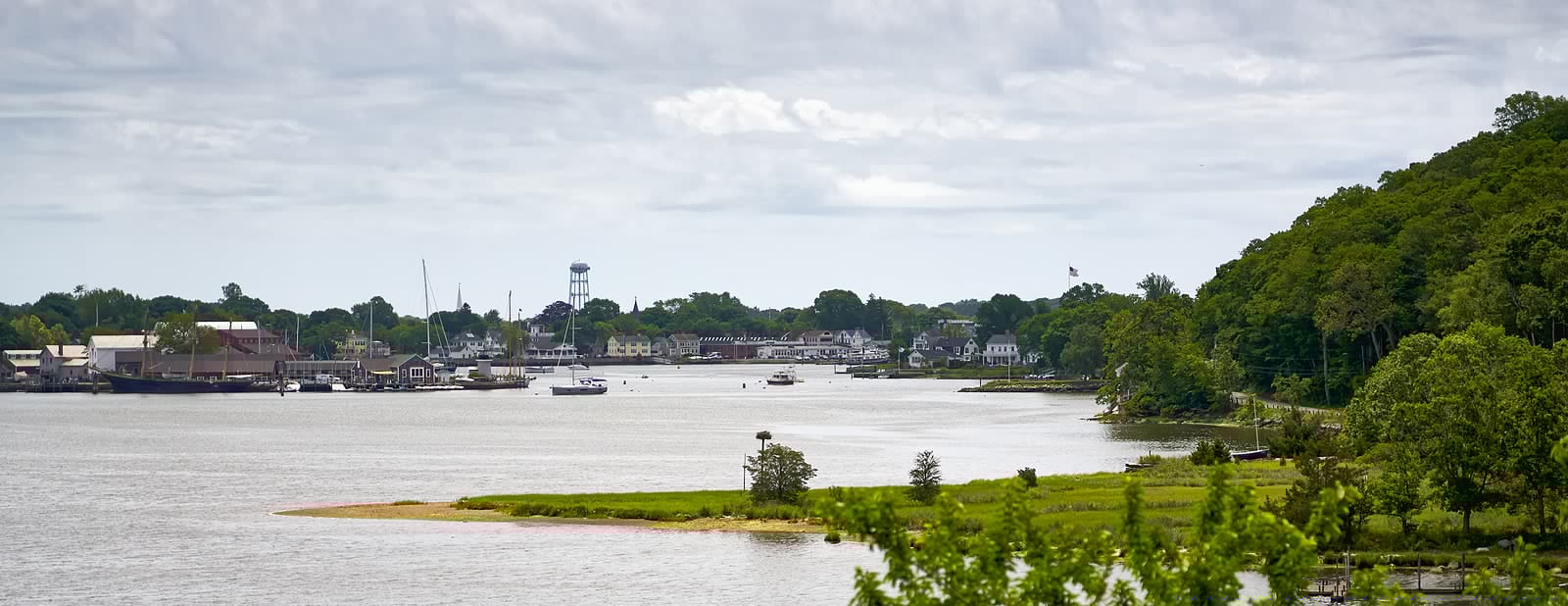 A scenic view of a river near a small town, surrounded by lush greenery and cloudy skies.