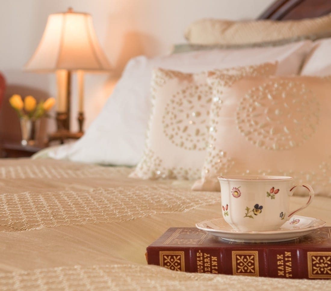 A delicate teacup rests on a stacked book atop a neatly made bed with decorative pillows and soft lighting in the background.