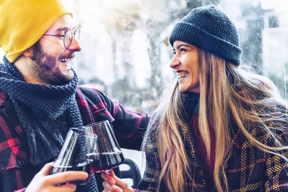 A couple wearing winter attire smiles at each other while toasting with glasses of red wine.