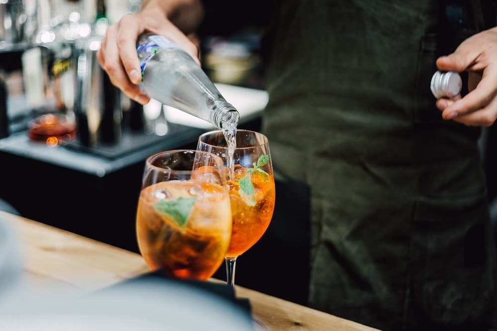 A bartender pours sparkling water into two cocktail glasses filled with ice and mint.