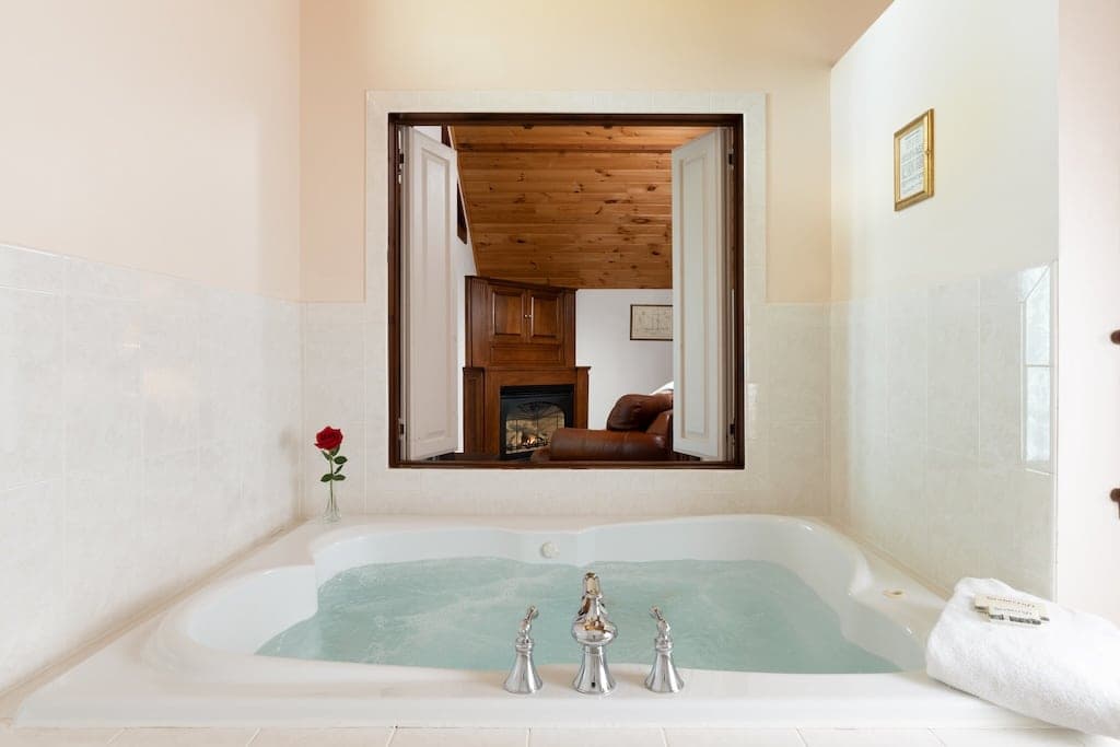 A luxurious whirlpool bath with a rose and a view of a cozy living area through an open window.