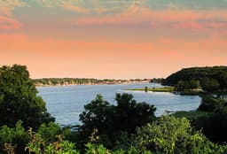 A serene river view bordered by lush greenery under a pastel sky at sunset.