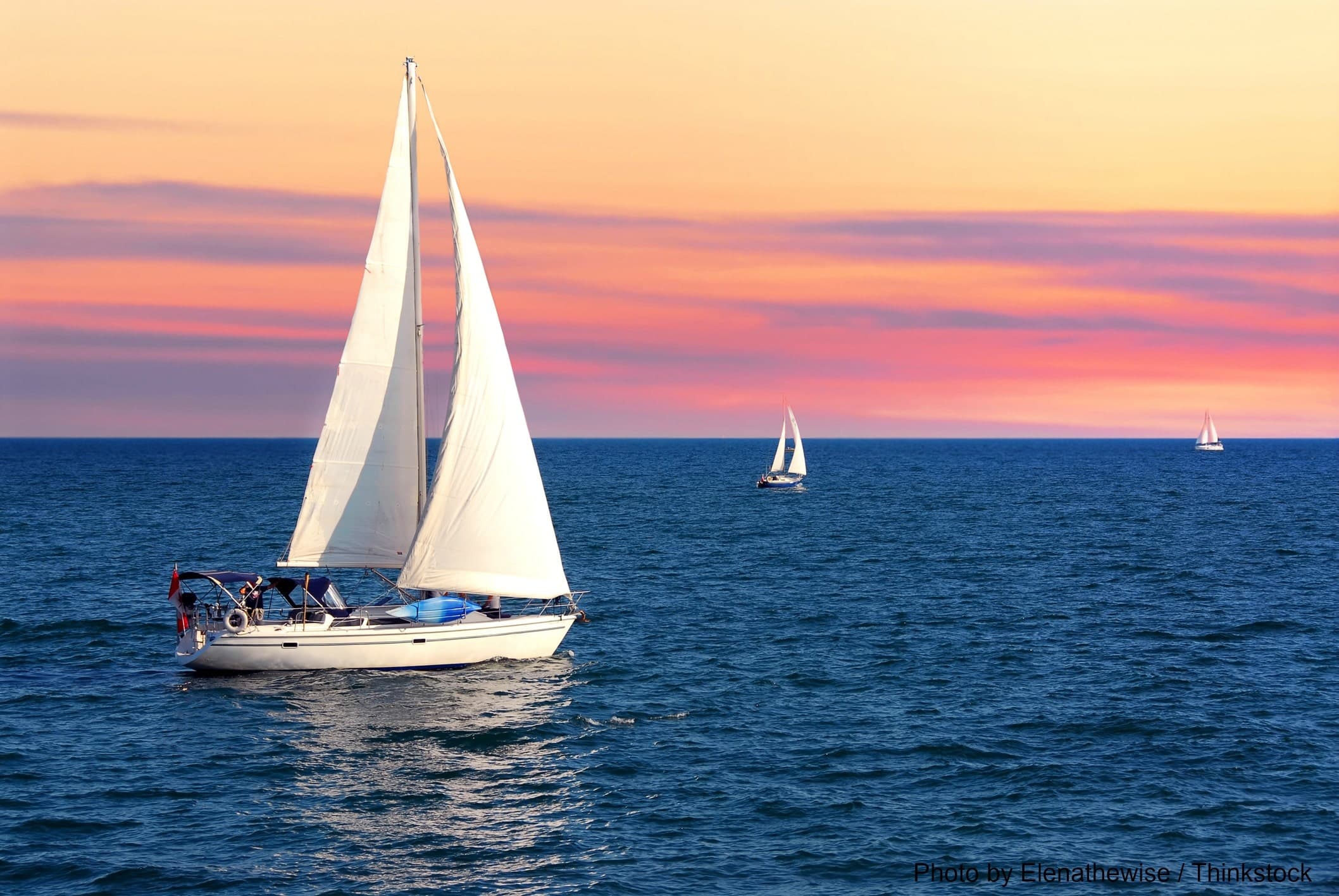 A sailboat glides across the water under a colorful sunset, with additional sailboats in the distance.