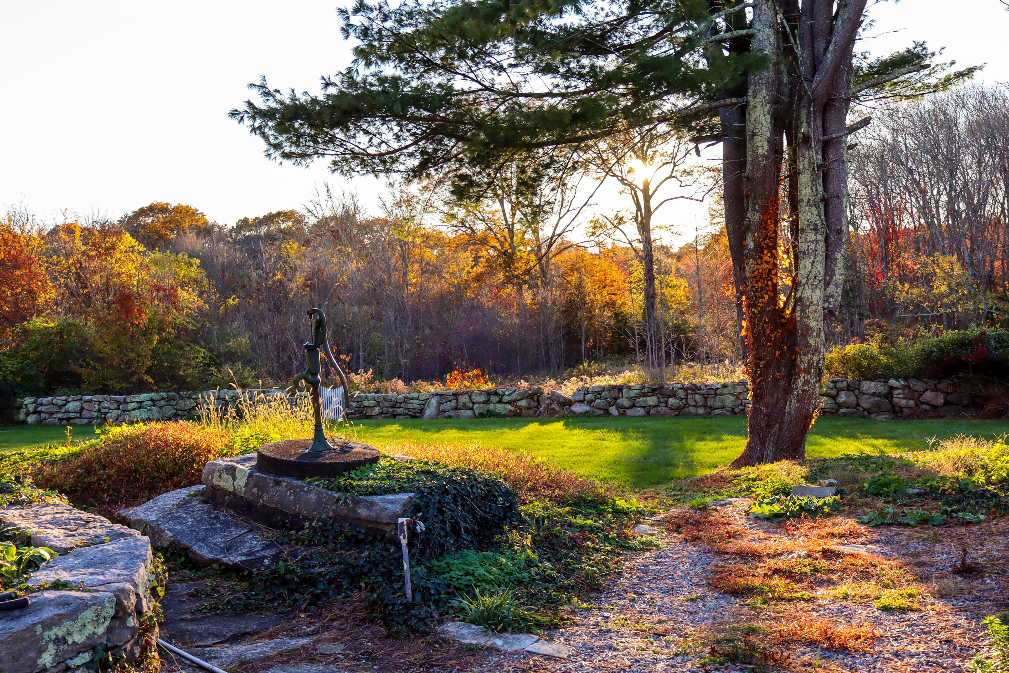 A sunlit landscape featuring a well surrounded by autumn foliage and stone walls.