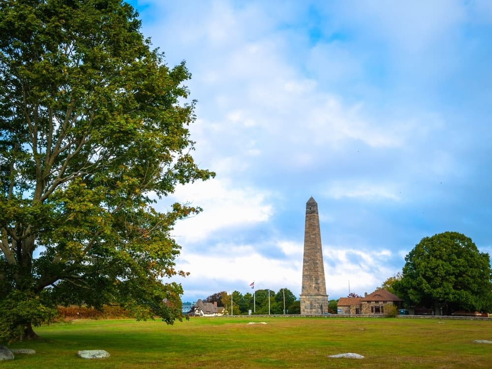 A tall stone monument stands in a grassy area surrounded by trees and buildings under a cloudy sky.