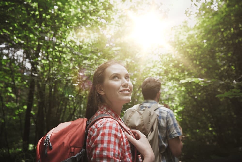 A woman smiles while hiking in a sunlit forest, with a man in the background. A woman smiles while hiking in a sunlit forest, with a man in the background.