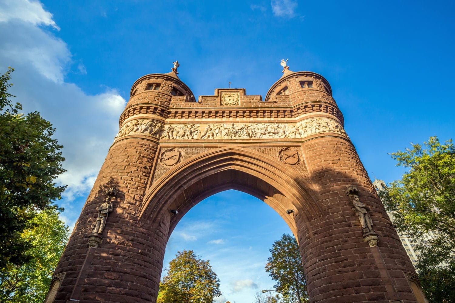 Red stone archway with towers under a blue sky, surrounded by trees.