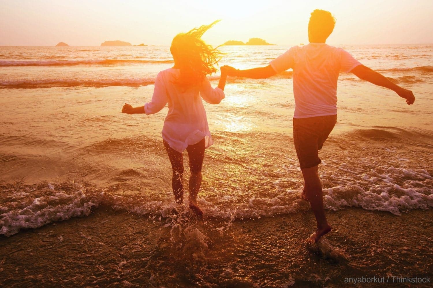 A couple runs hand in hand through the shallow waves at sunset.