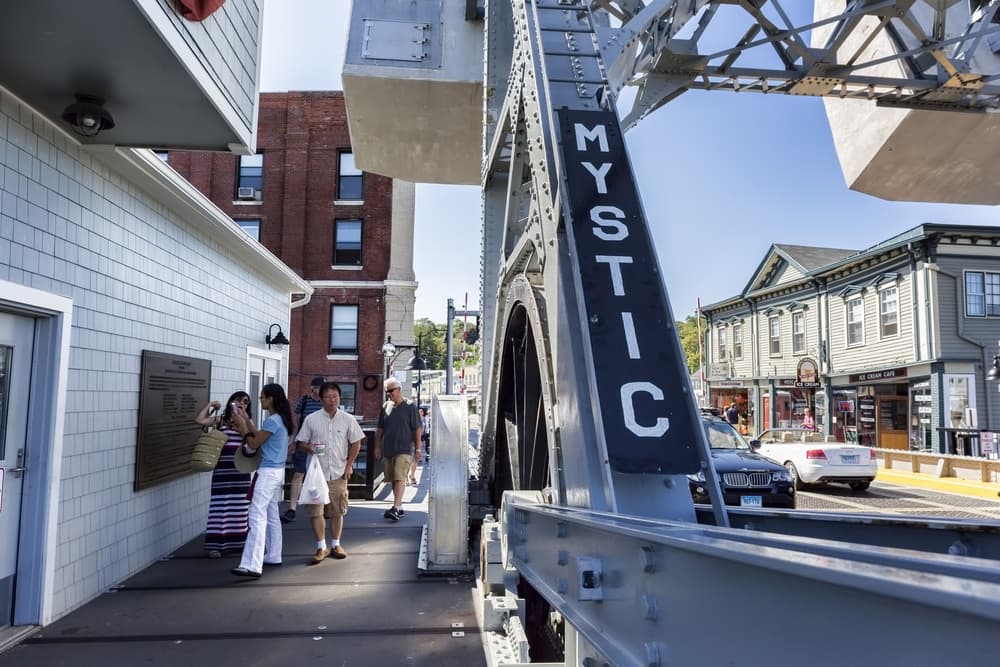 People walk by a bridge structure marked "MYSTIC" in a quaint coastal town.