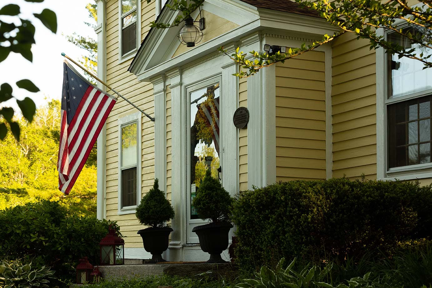 A yellow house with an American flag, a wreath on the door, and neatly trimmed shrubs.