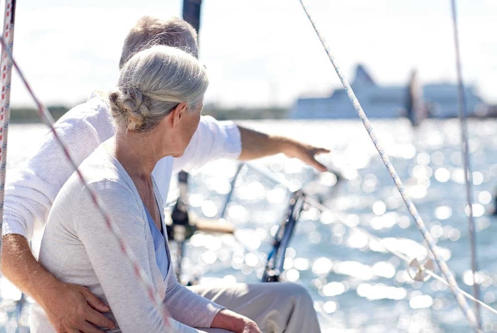 An older couple enjoys a sailboat ride, with one pointing towards the shimmering water.