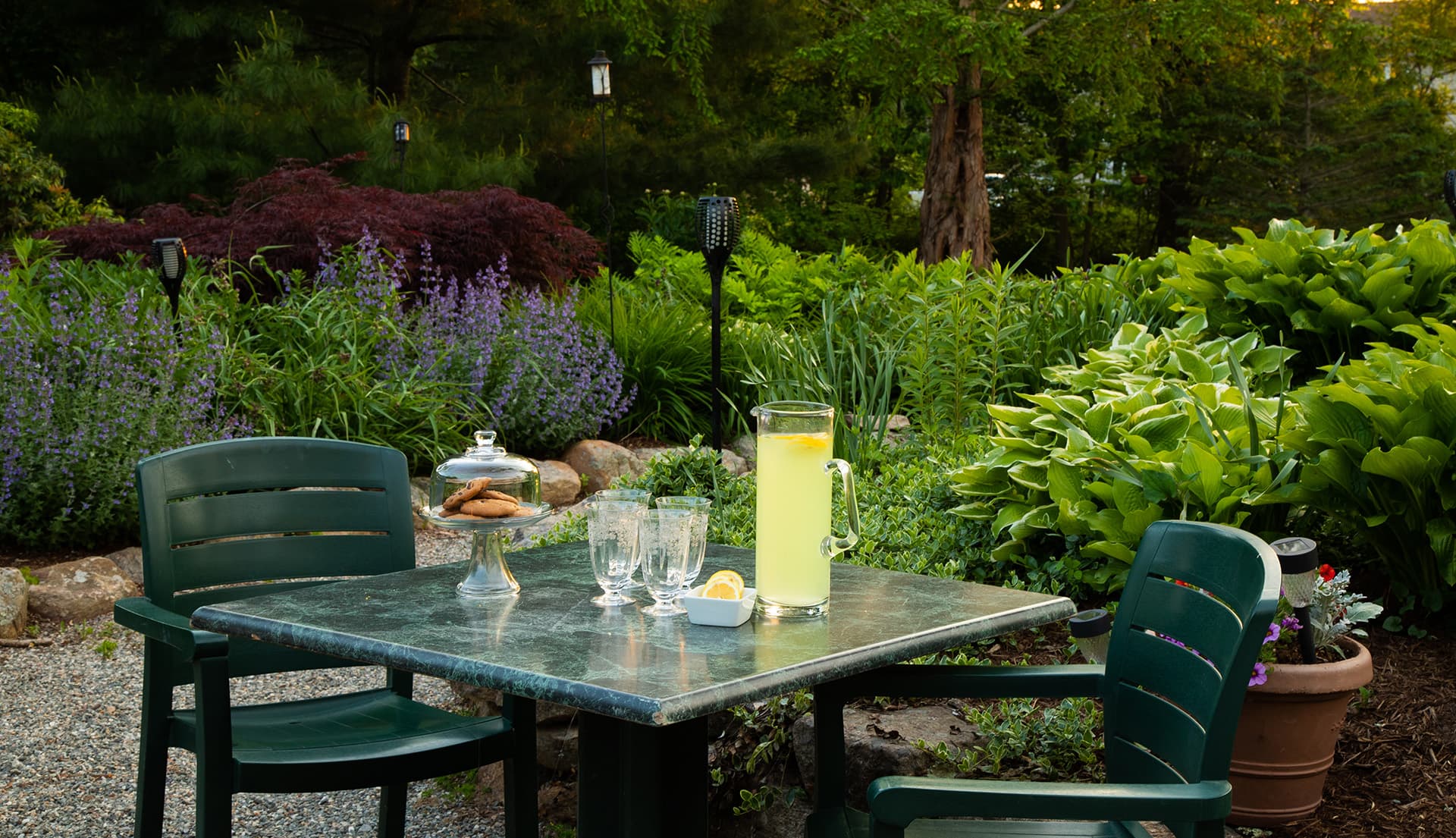 A table with two chairs set in a lush garden, featuring a pitcher of lemonade and a glass dish of cookies.
