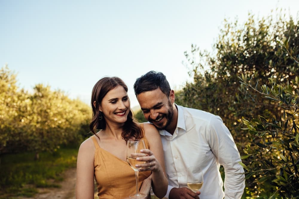 A couple joyfully shares a moment in an olive grove while holding glasses of wine.