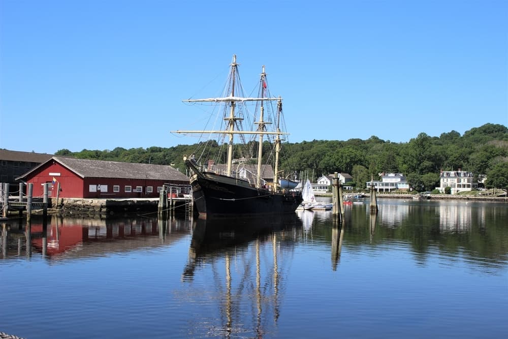 A tall ship docked in calm waters with reflections of nearby buildings and trees.