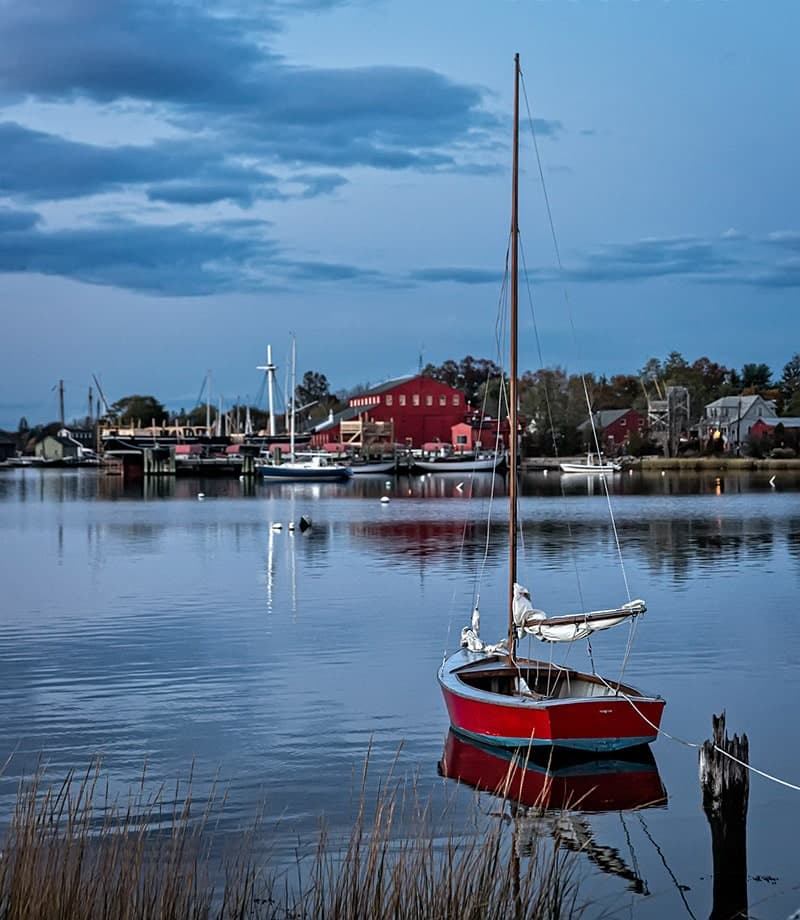 A red sailboat is anchored in calm water with a scenic waterfront and cloudy sky in the background.