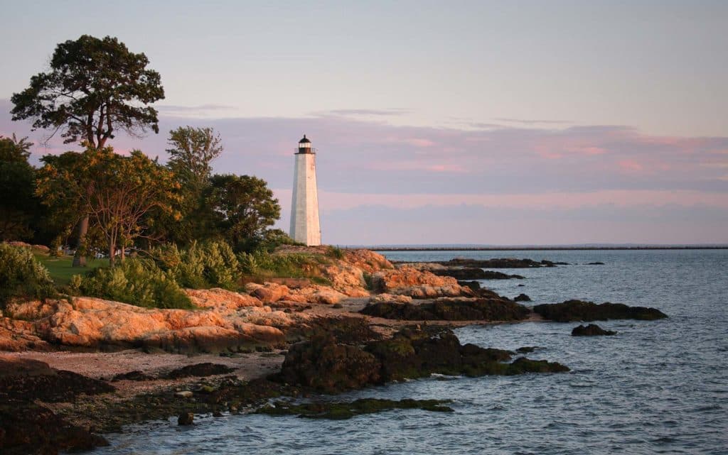 A lighthouse stands by the coastline at sunset, surrounded by trees and rocky shoreline.