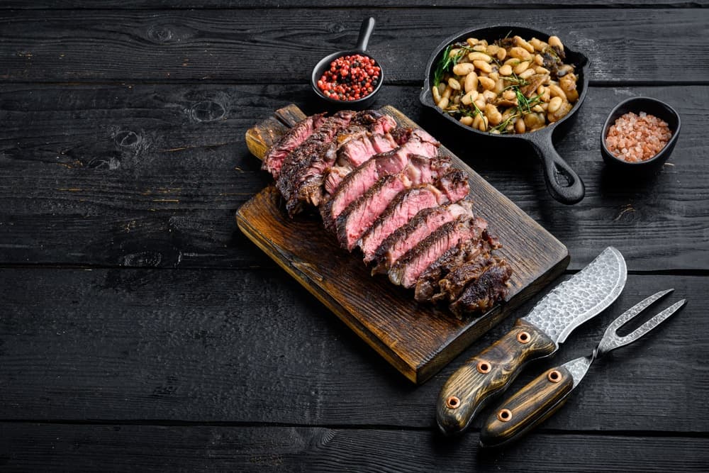 Sliced steak on a wooden cutting board with sides of beans and pink peppercorns.