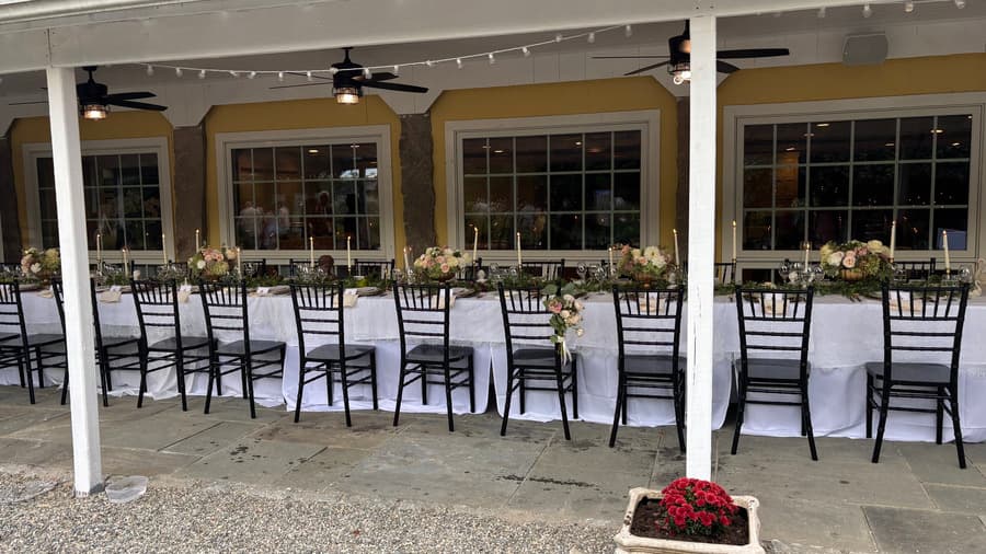 A long, elegantly set dining table with black chairs and floral centerpieces, under a covered patio.