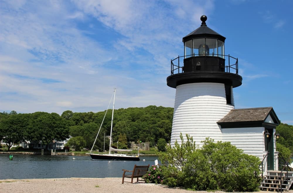 A white lighthouse stands by the water, with a sailboat in the background and lush greenery surrounding it.