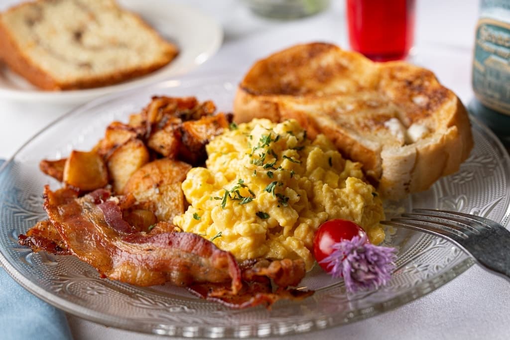 A plate of scrambled eggs, crispy bacon, roasted potatoes, a slice of toast, and a cherry tomato garnish.