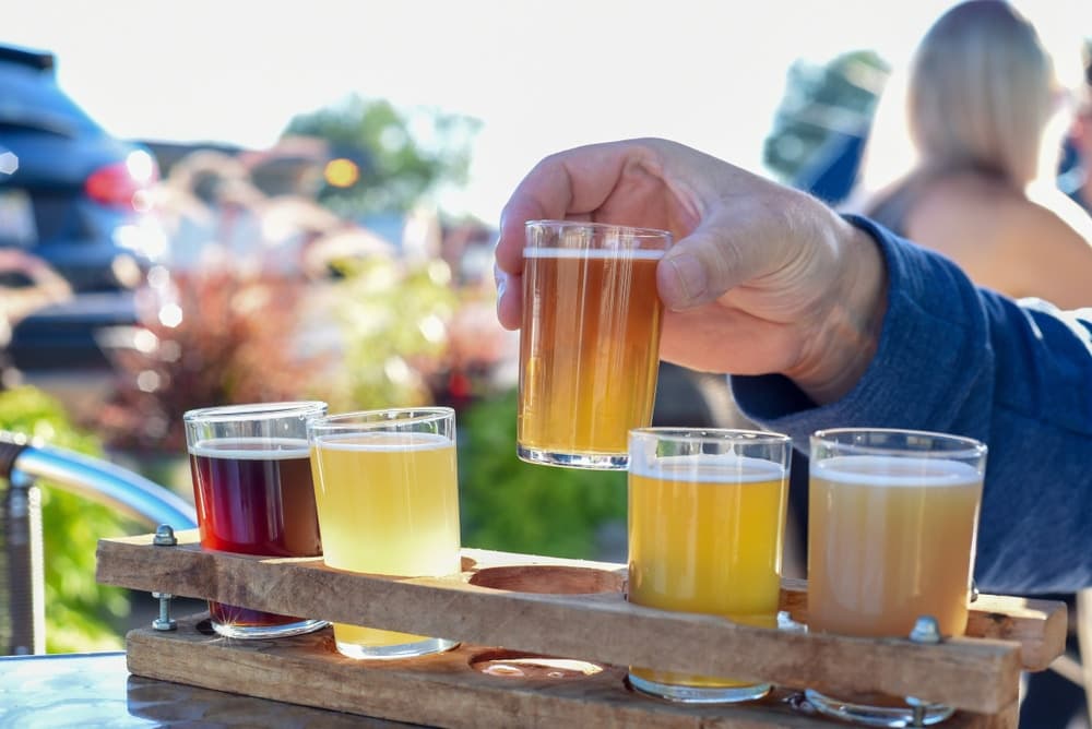 A hand holds a small glass of beer above a wooden flight with various beer samples.