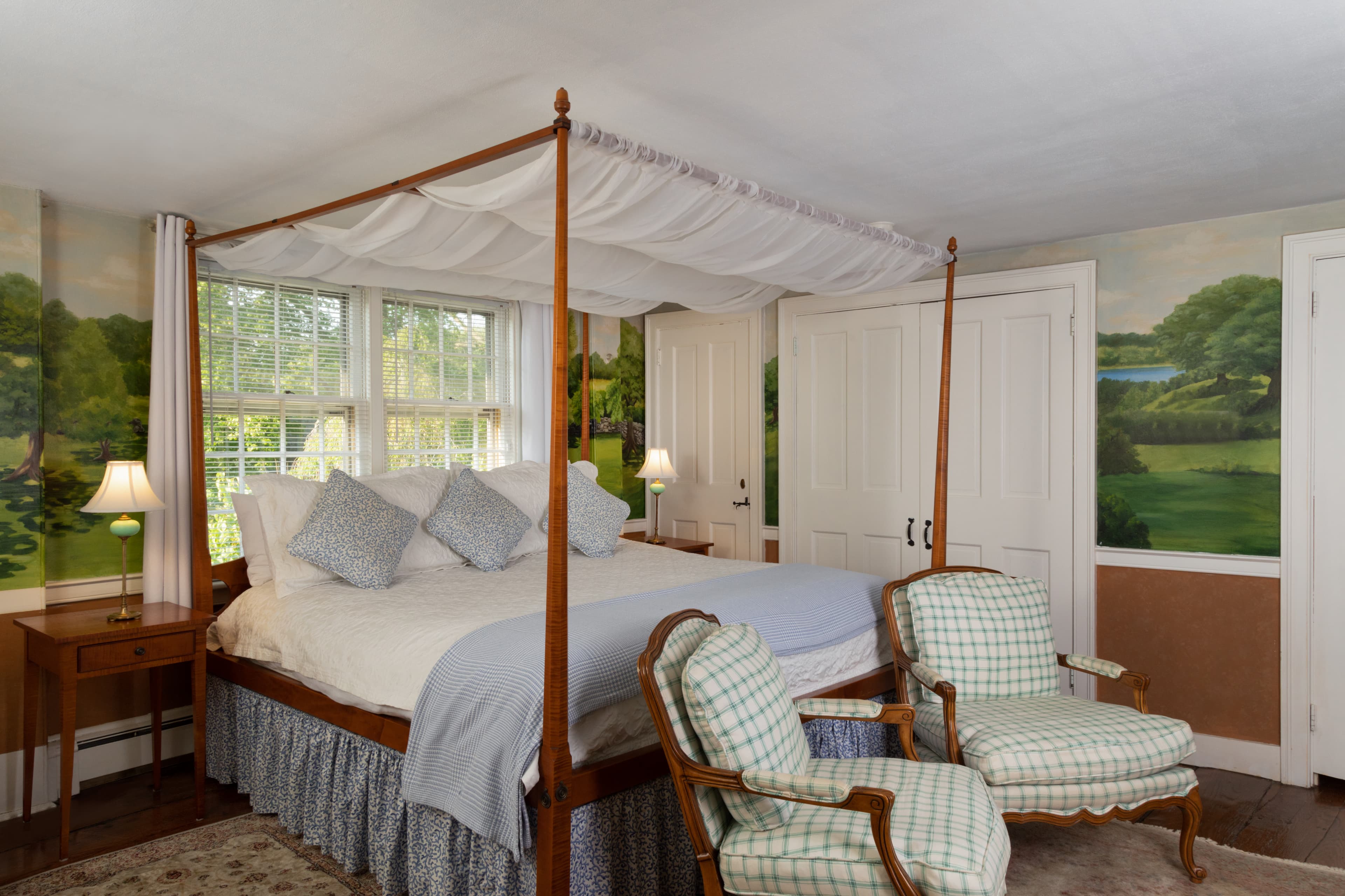 A cozy bedroom featuring a four-poster bed, patterned chairs, and serene landscape murals.