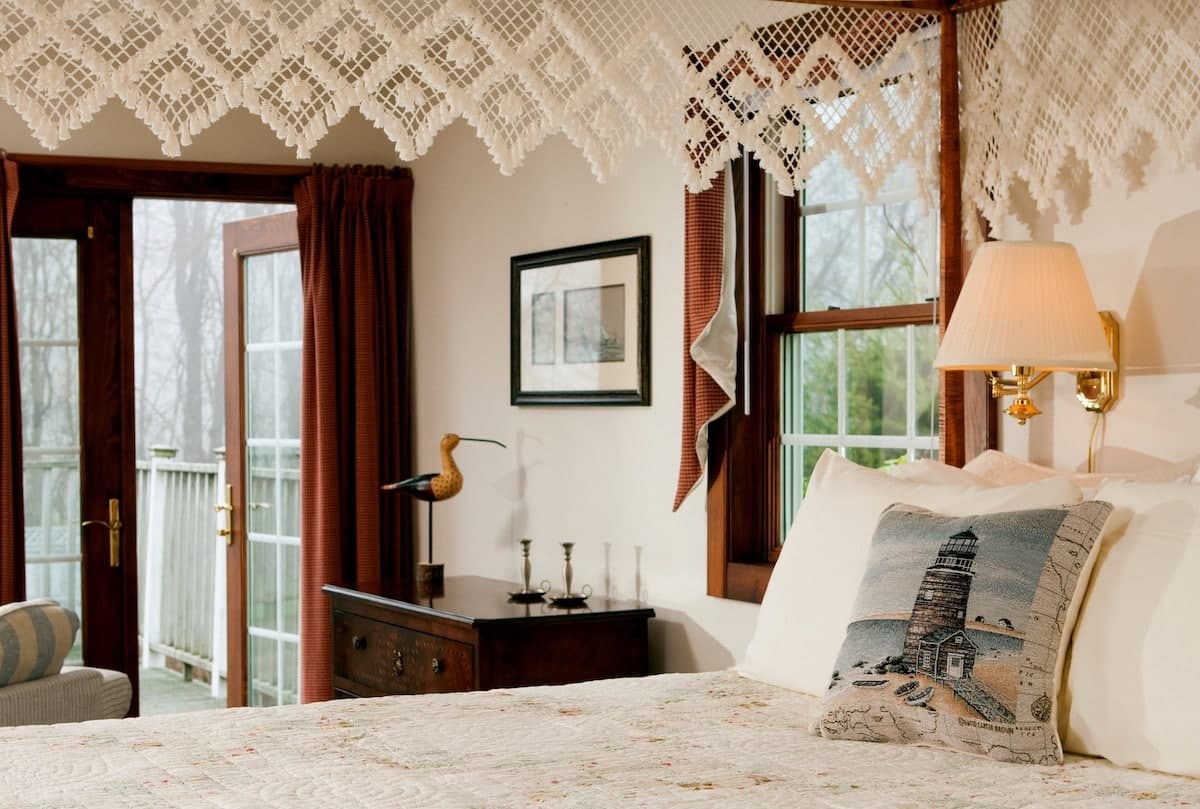 A cozy bedroom with a lace curtain, framed artwork, and a bedside lamp, featuring a view of a balcony through double doors.