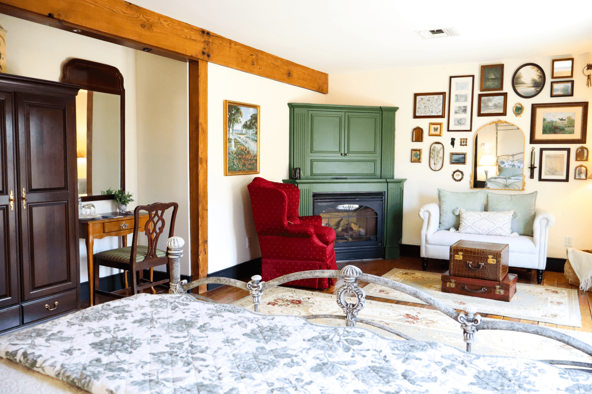 A cozy bedroom featuring a floral-patterned bedspread, an upholstered chair, a green cabinet, and various framed pictures on the walls.