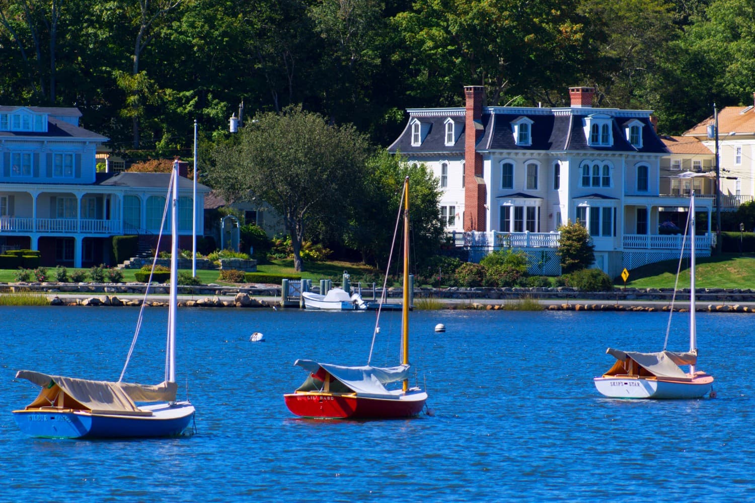 Four small sailboats are anchored in a calm bay, with charming houses and trees in the background.