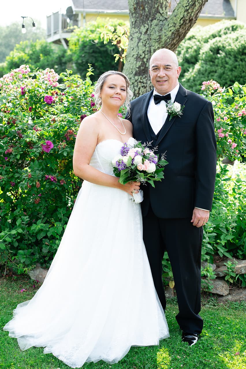 Bride in a white gown holding a bouquet stands beside a groom in a tuxedo, surrounded by lush greenery and flowers.